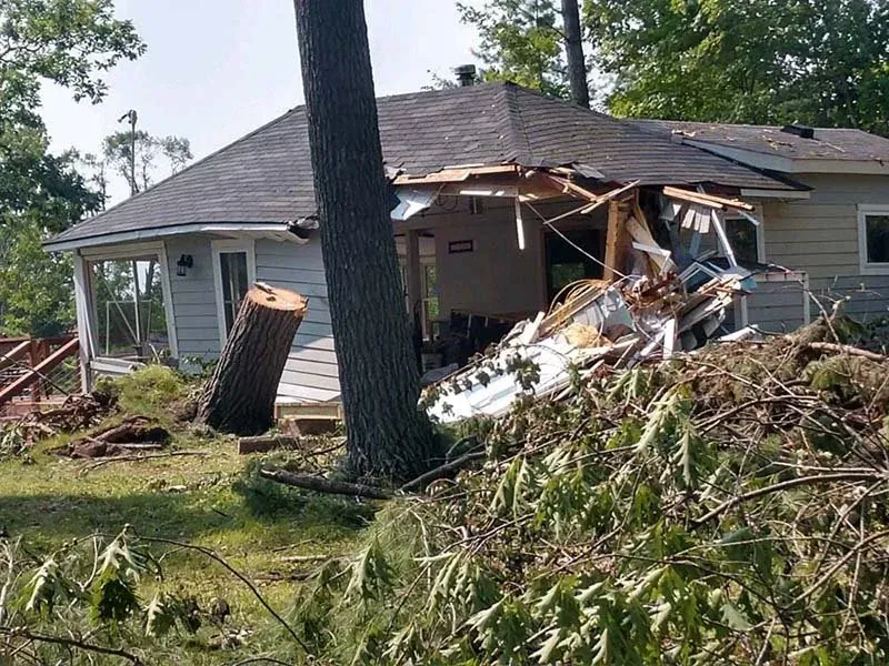 A house with significant storm damage, featuring a collapsed roof and debris, with a tree trunk nearby.