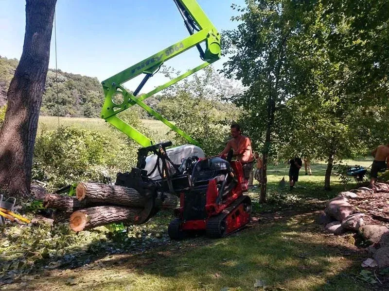 A person operates a red compact track loader with a grapple attachment lifting logs beneath a bright green aerial lift arm.