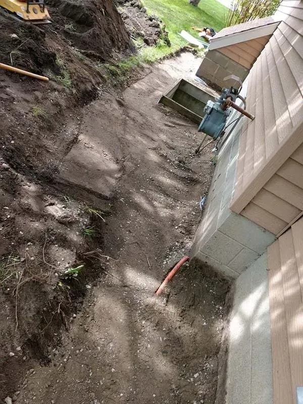 A narrow, excavated dirt path along the side of a house, featuring a gas meter and exposed pipes near the foundation.