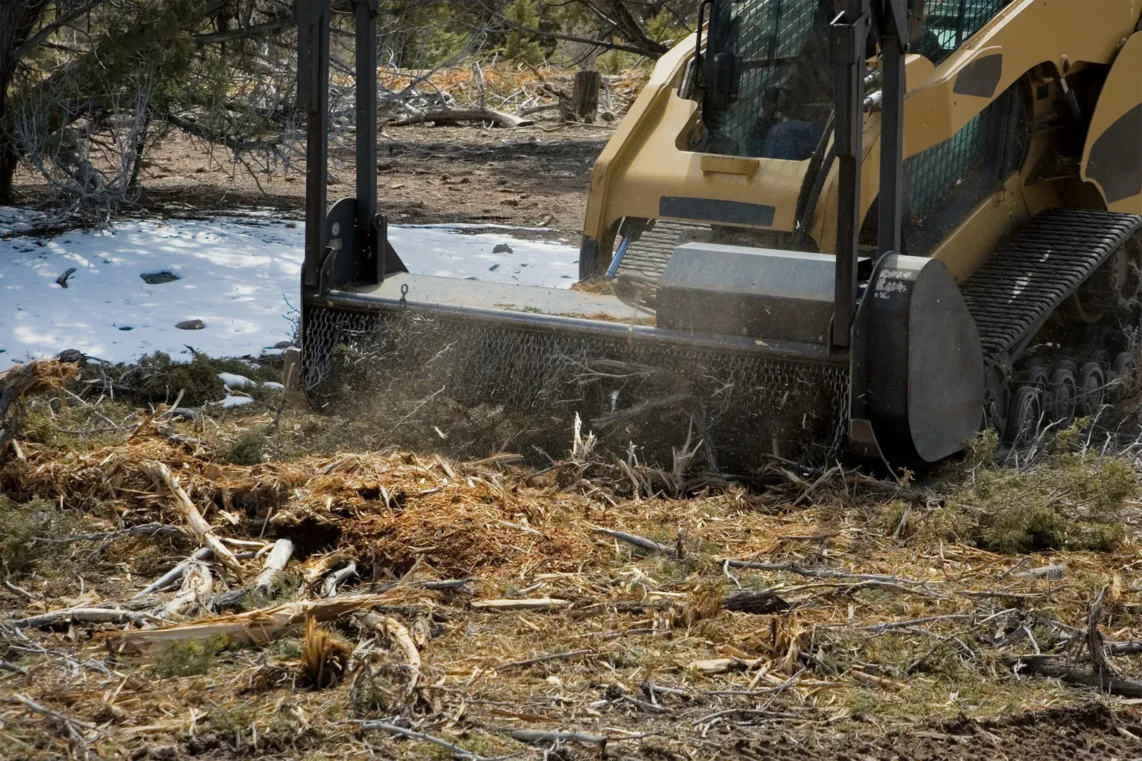 A yellow skid steer with a forestry mulcher attachment grinds brush on snowy ground.
