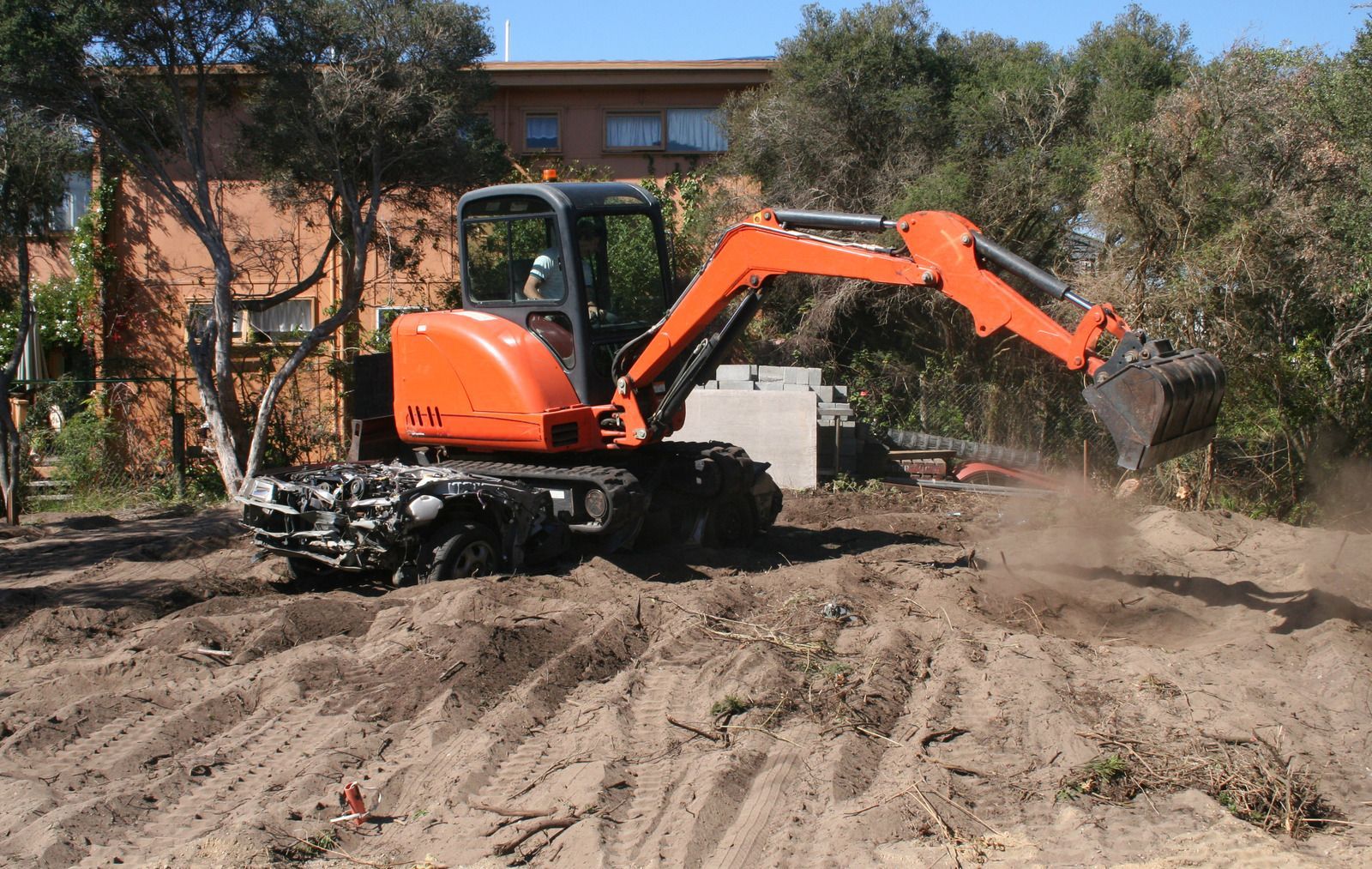 An orange excavator digs into dirt in an outdoor area near trees and a building.