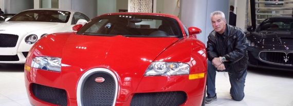 Red sports car in a showroom with a man crouching beside it; another black car is behind.