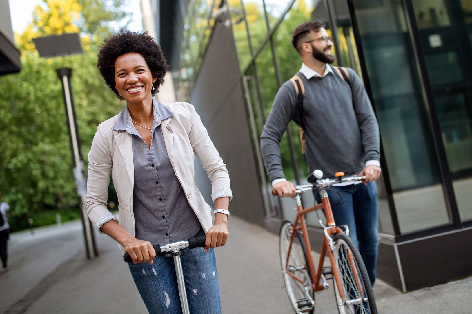 Woman on scooter smiles near a man on a bike; both ride near a modern building with trees in the background.