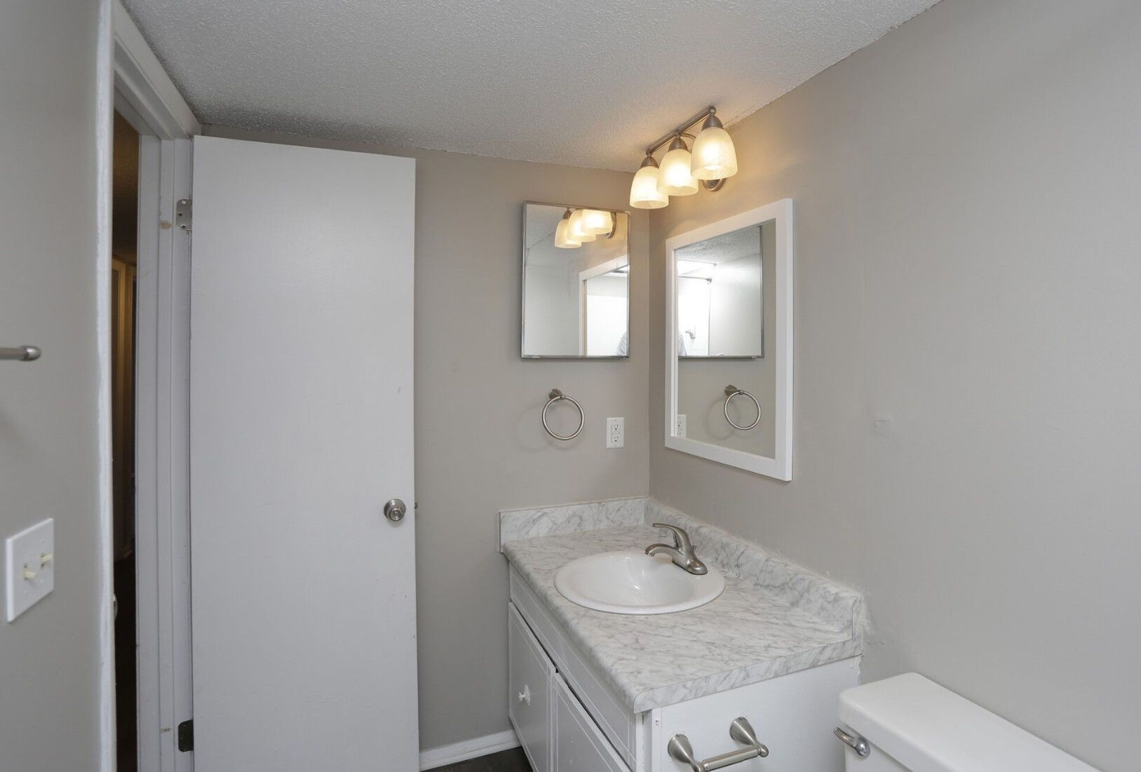 A small bathroom with a white vanity, mirror, and toilet. The walls are gray, and there's a partially open white door on the left.