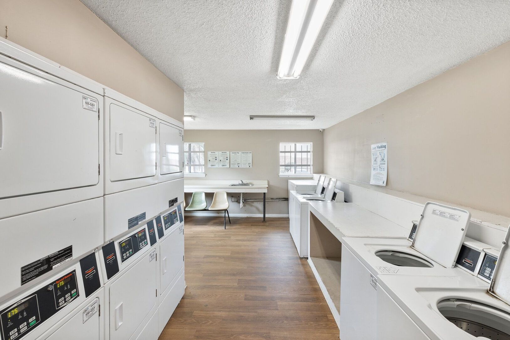 A brightly lit laundry room with rows of white washing machines and dryers, a sink, and a long counter.