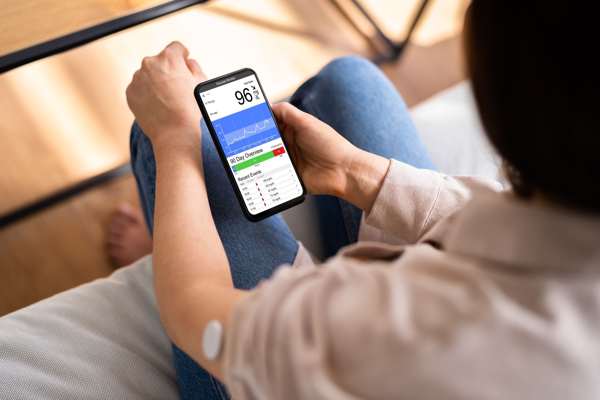 Person sitting, holding phone displaying blood sugar data; sensor on arm.