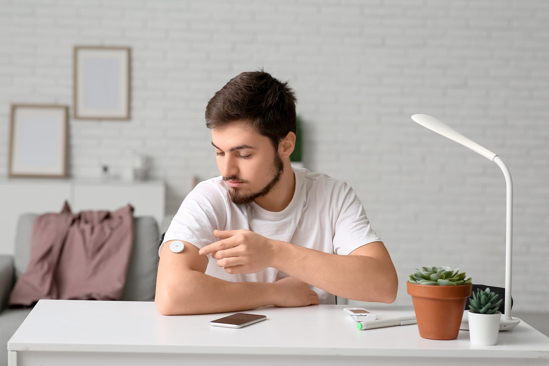 Man with a white t-shirt checking a sensor on his arm, sitting at a desk, with a plant and lamp.