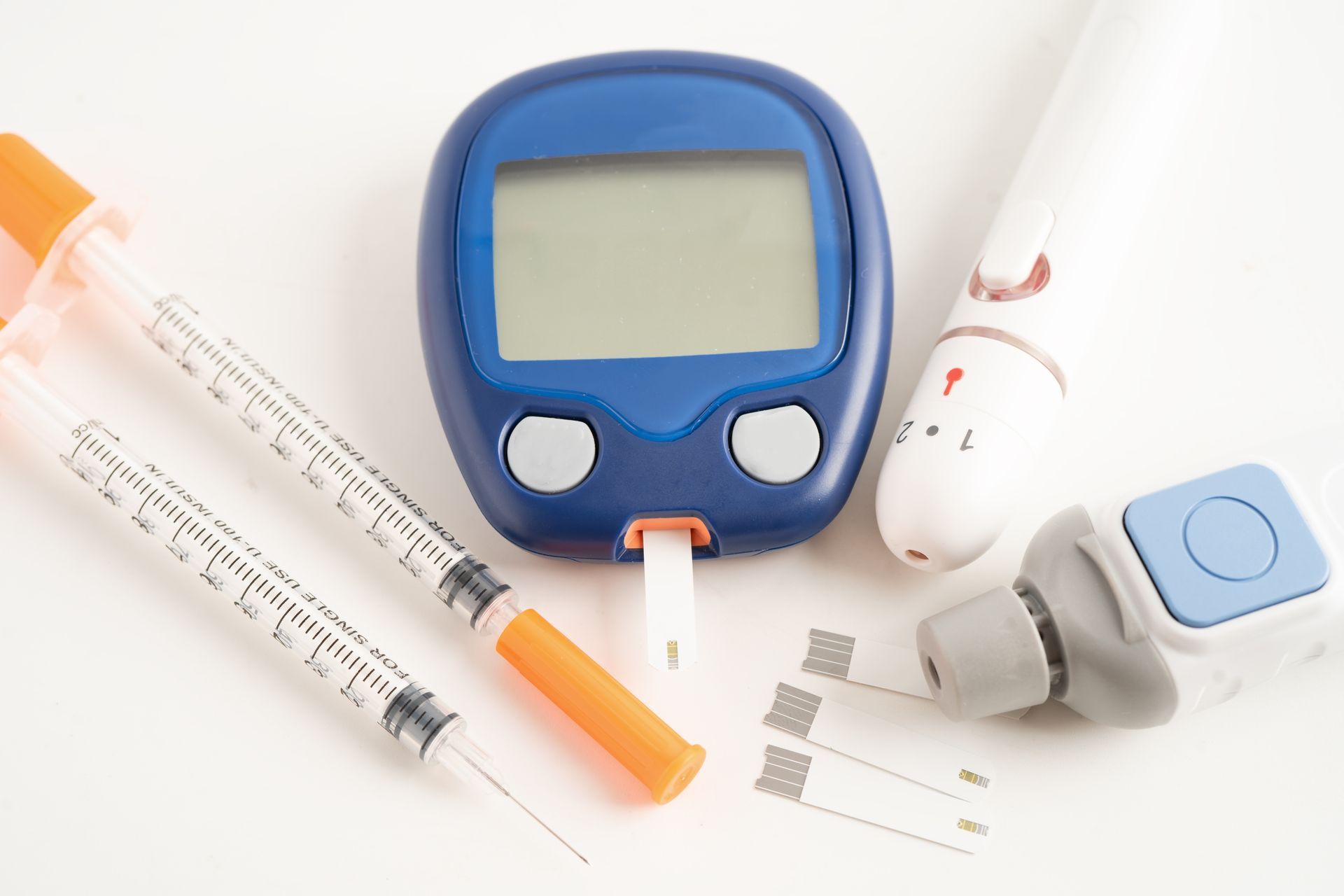 Diabetes management tools: glucose meter, lancet, insulin syringes, and test strips on a white surface.