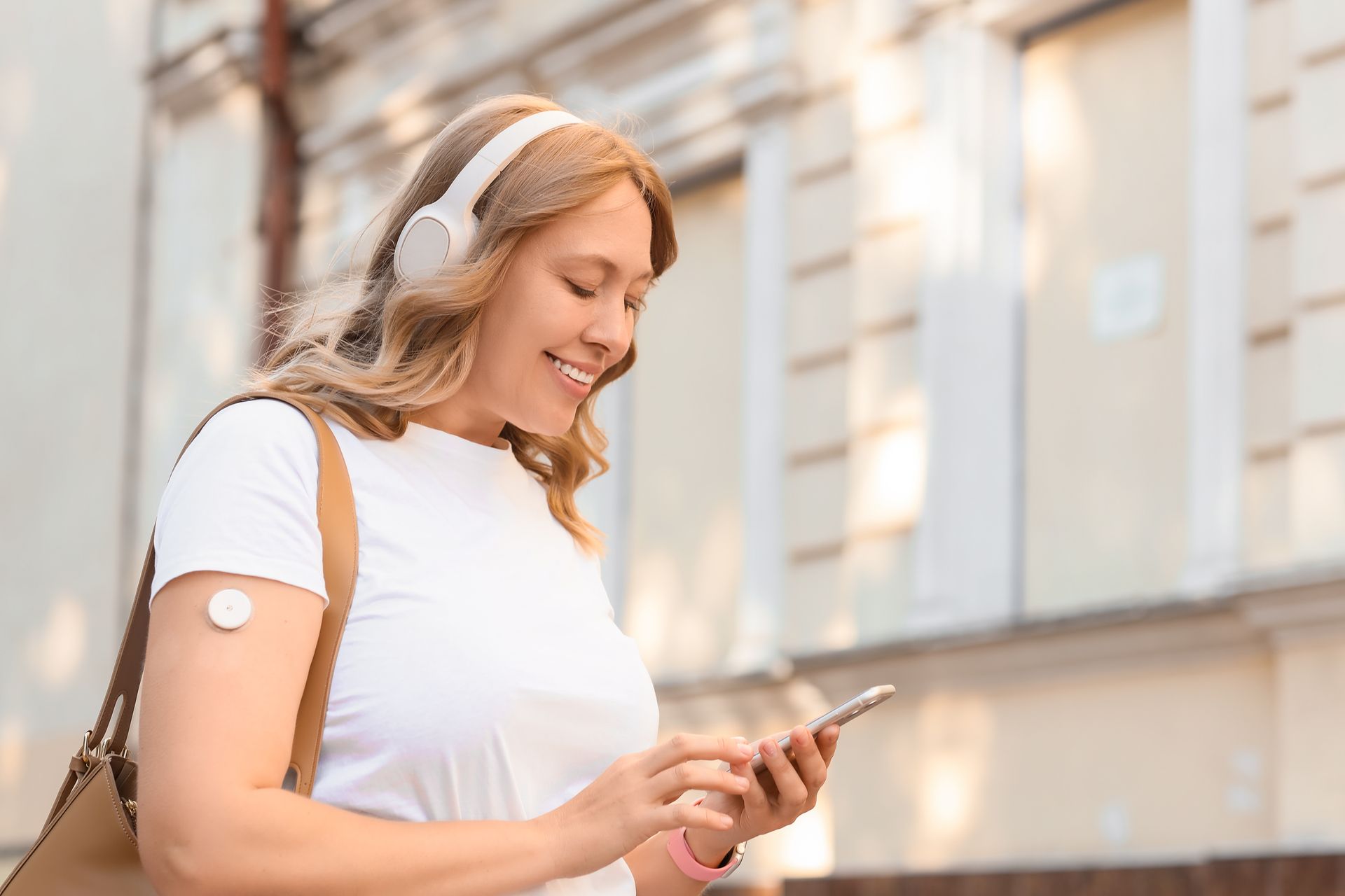 Woman smiling, wearing headphones, checking phone, glucose monitor on arm, outdoors.