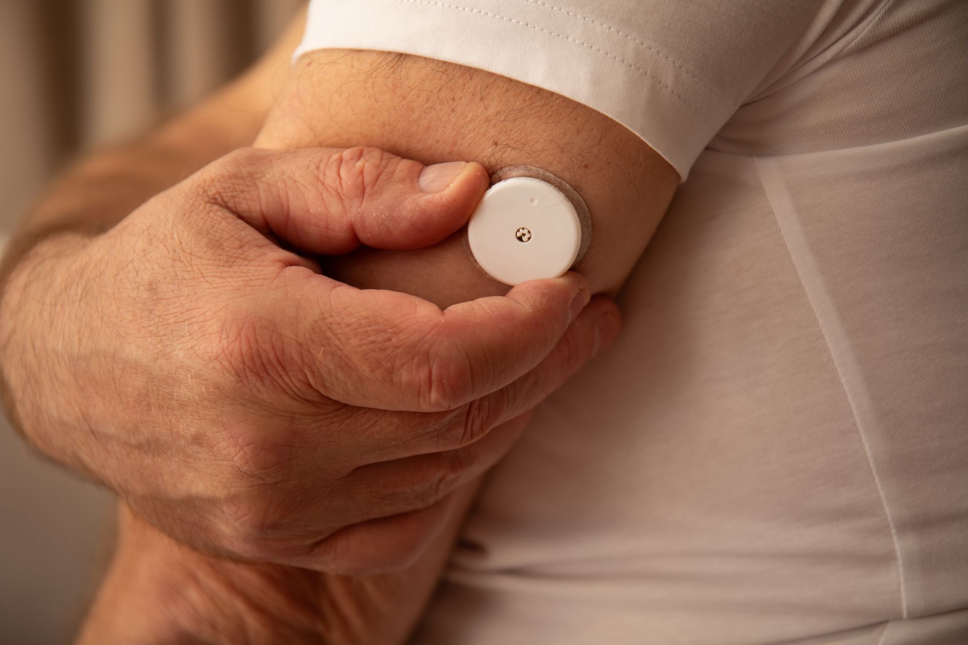 Person's arm with a round, white medical sensor attached. Hand is holding the sensor. White shirt.