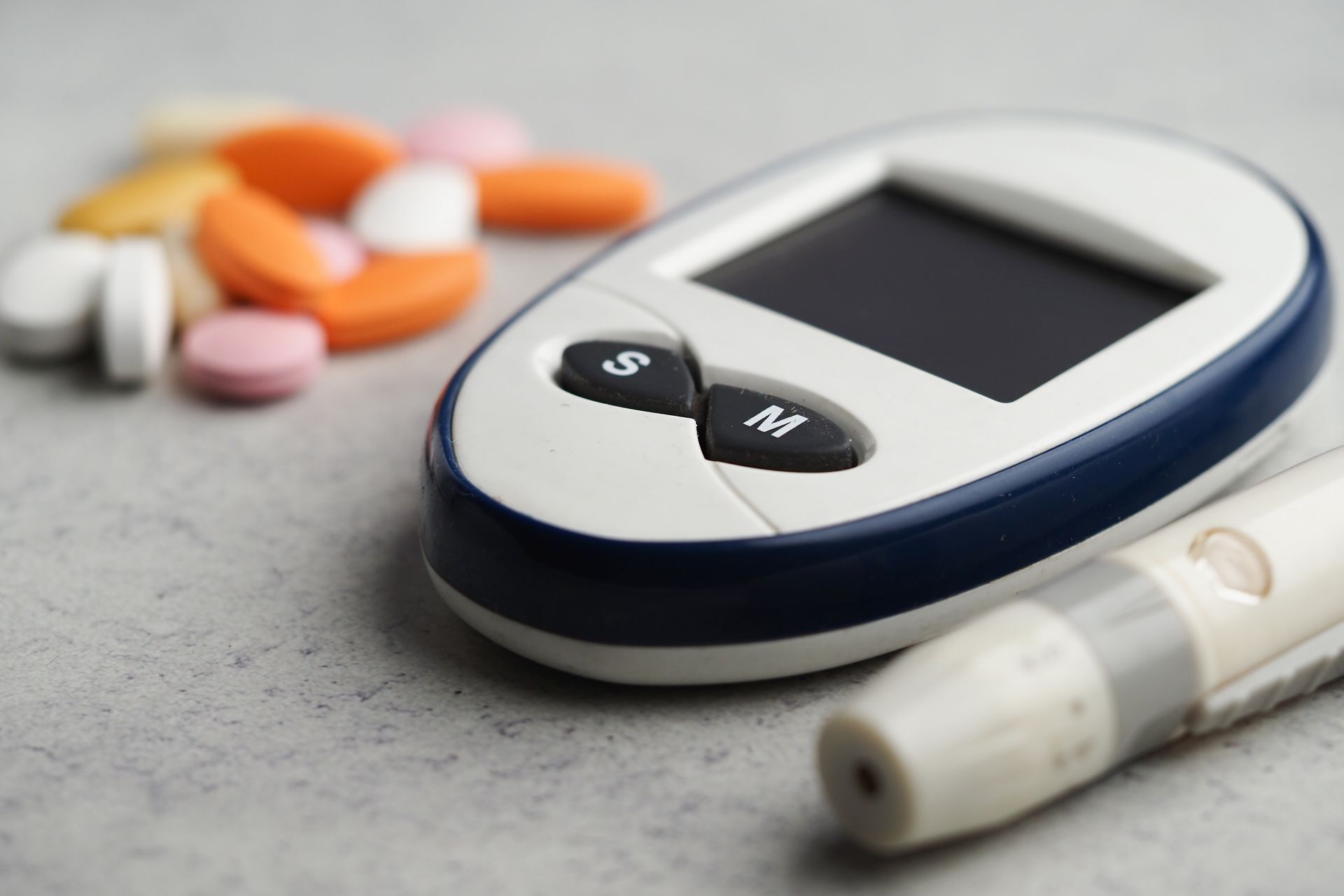 Diabetes supplies, including pills, blood glucose monitor, and lancing device on a grey surface.