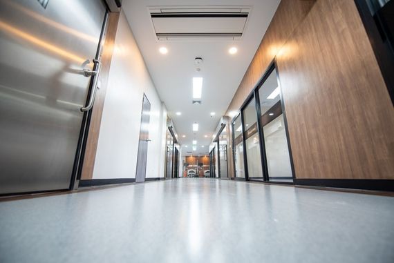 A long, empty indoor hallway with a polished floor, a metal door on the left, and glass-paneled walls on the right.
