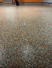 A person uses a roller to apply a gray, black, and orange speckled epoxy coating to a floor.