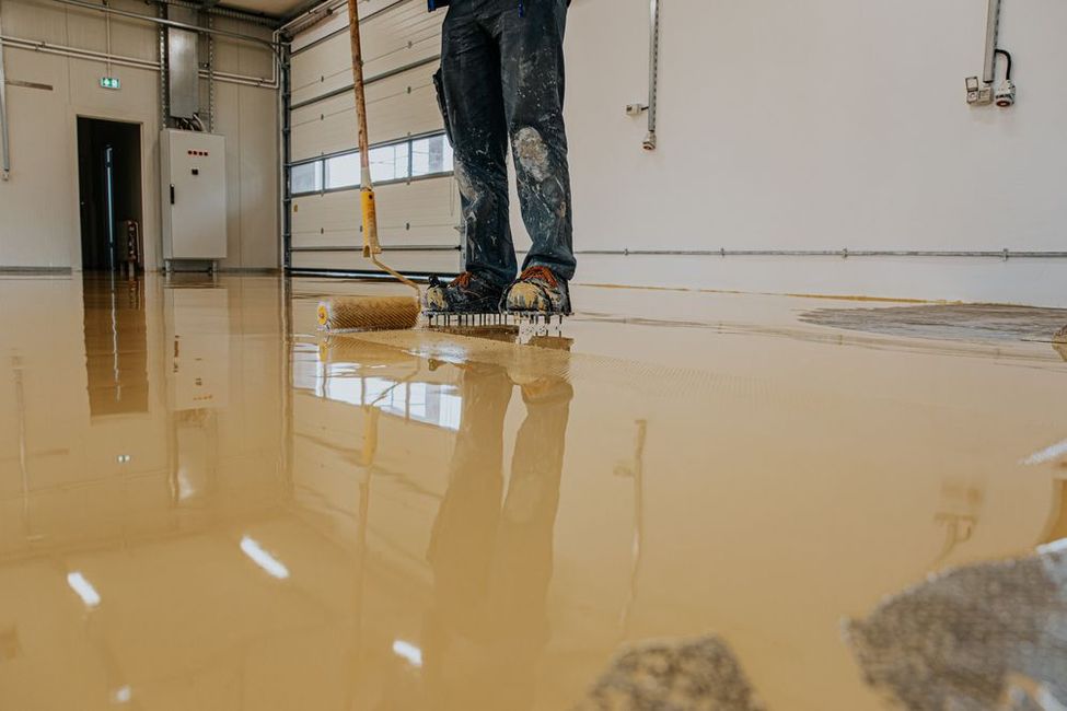A worker wearing spiked shoes uses a roller to apply smooth, light brown epoxy coating to a warehouse floor.