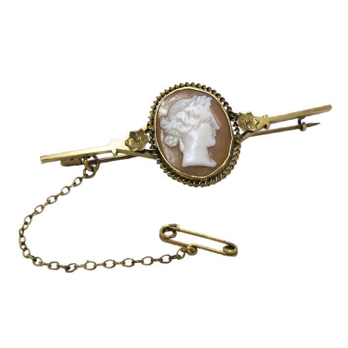 A gold bar brooch featuring a central oval shell cameo of a profile portrait, attached to a safety pin by a thin chain.