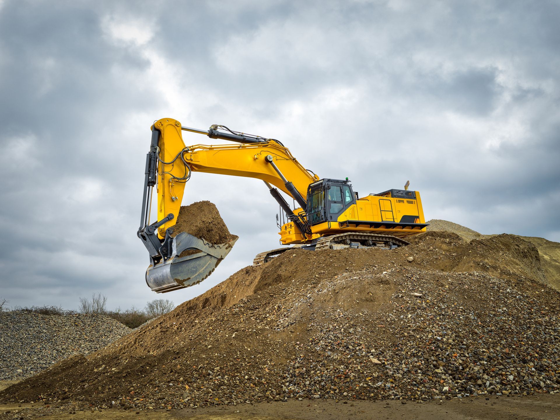 Yellow excavator digging into a pile of dirt under a cloudy sky.