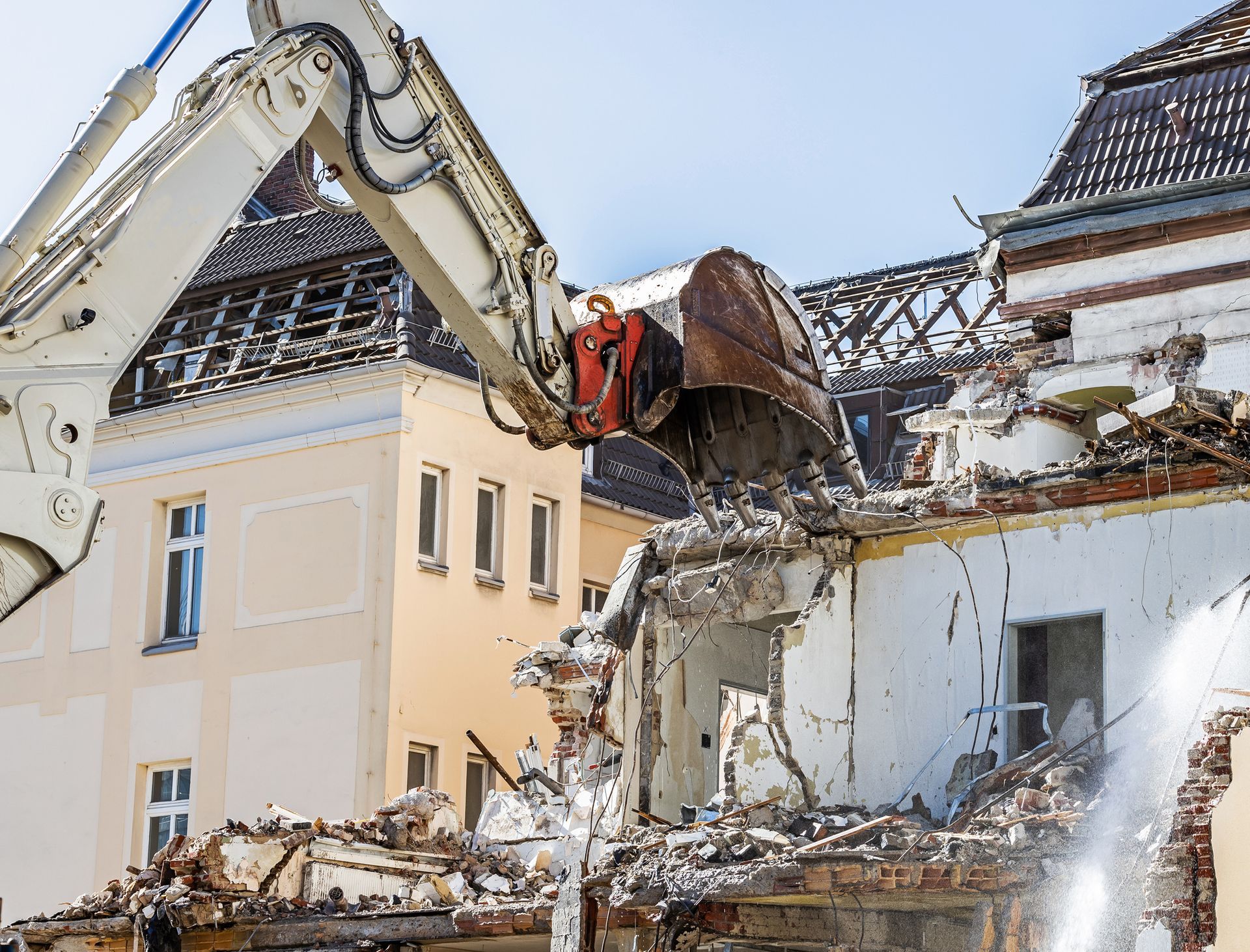 Demolition of a building with an excavator; beige and white buildings, blue sky.