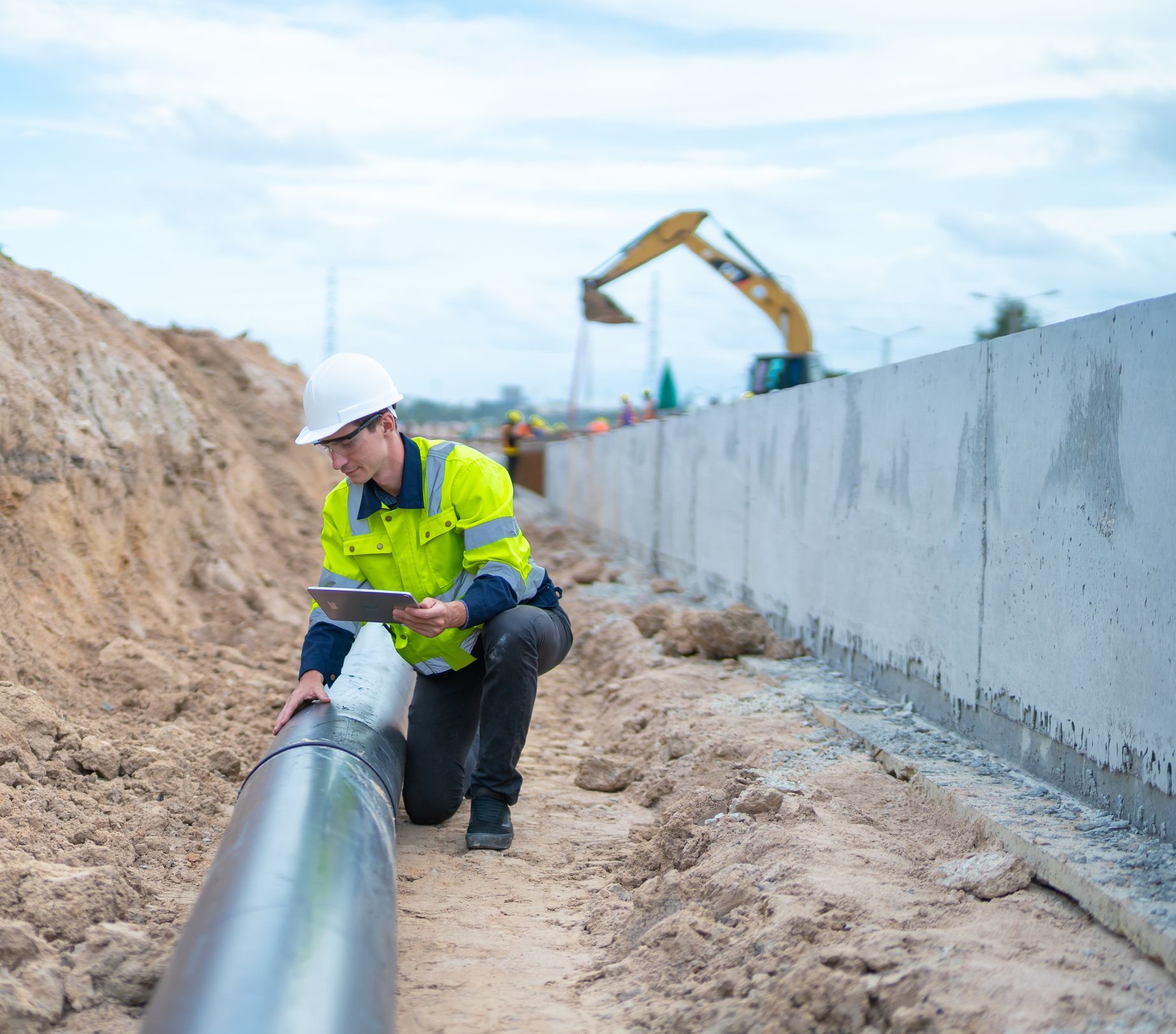 Construction worker inspecting a pipe in a trench, using a tablet, wearing a helmet and reflective vest, with a construction site in the background.