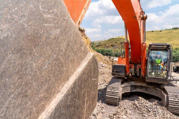 Orange excavator bucket scoops dirt, operator in the cab, outdoors.