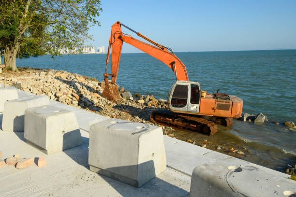 Orange excavator working on a concrete and rock seawall next to the ocean.