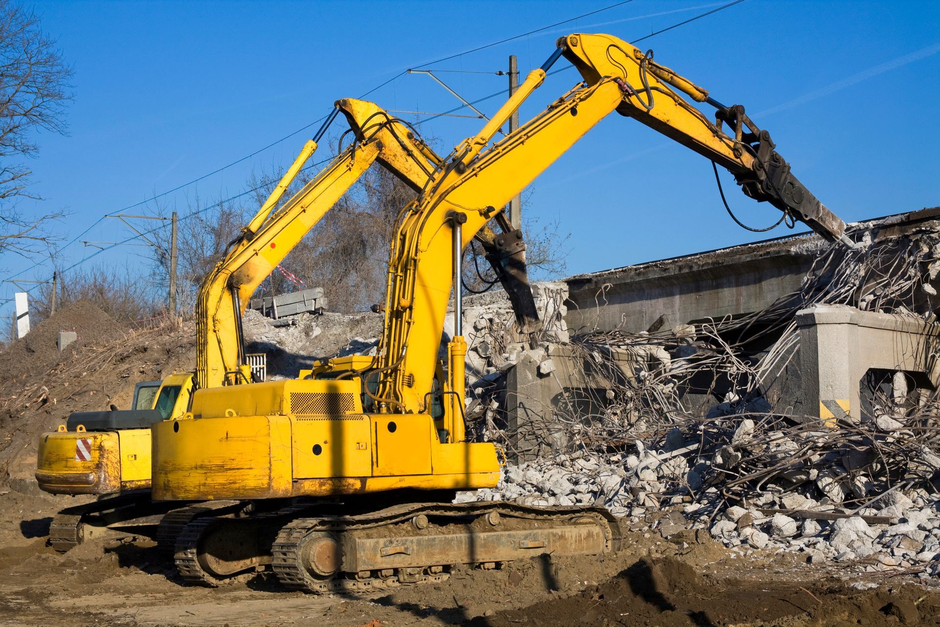 Two yellow excavators demolish a concrete structure on a sunny day.