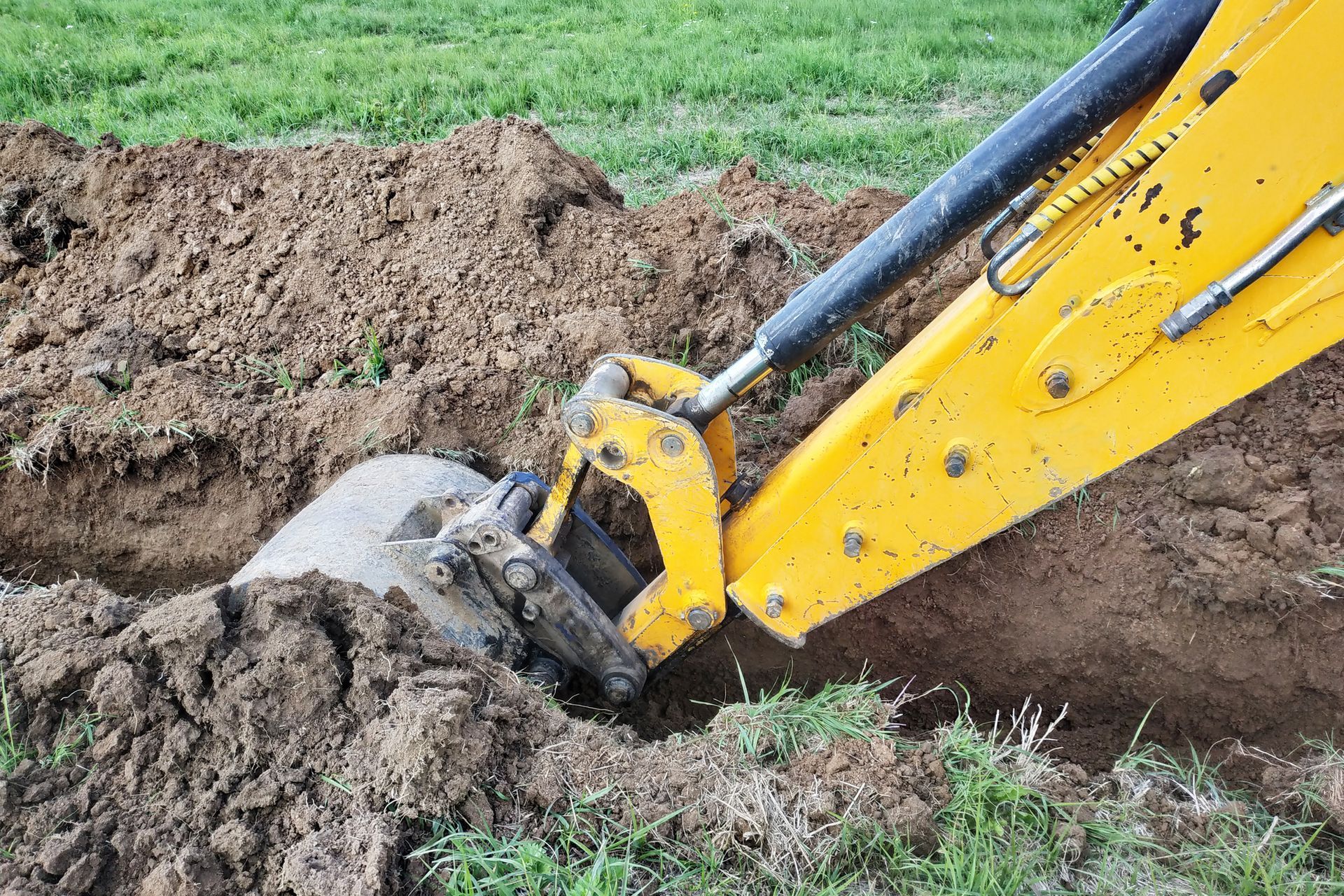 A yellow backhoe excavates a trench in a grassy field, near a large, gray rock.