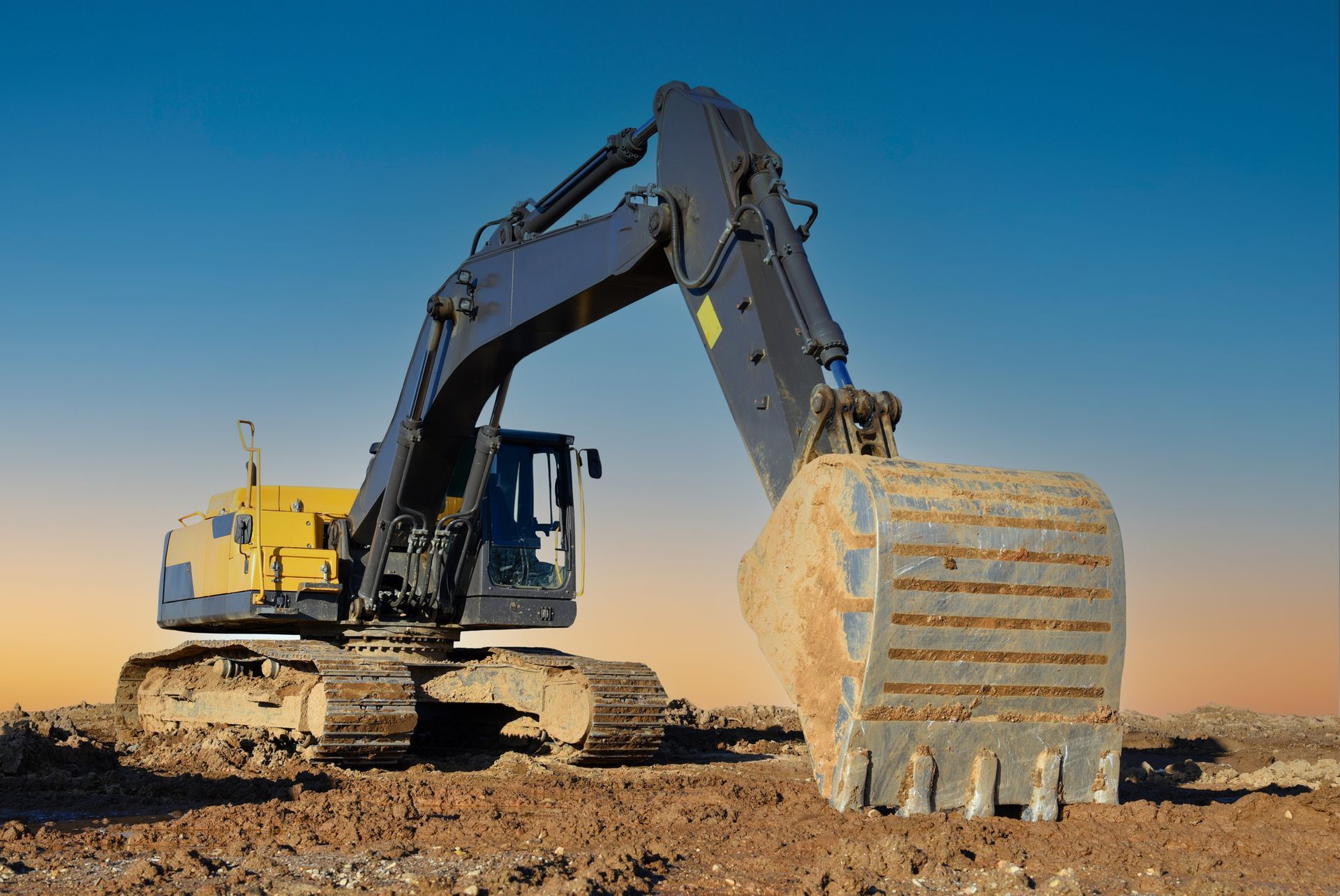 Yellow and blue excavator digging in a dirt field with a blue and orange sky.