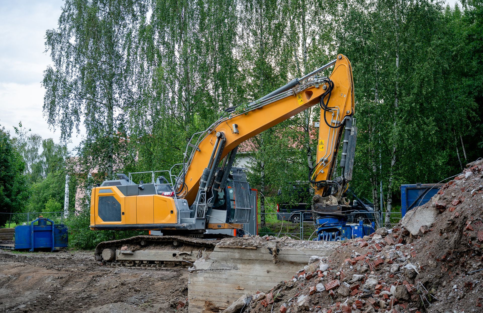 Yellow excavator demolishing a concrete structure, surrounded by dirt and rubble. Green trees are in the background.