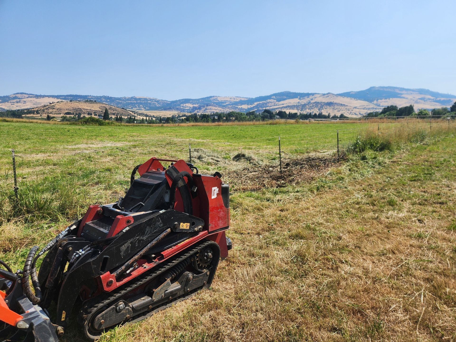 Mini excavator in a field with a distant mountain range under a blue sky.