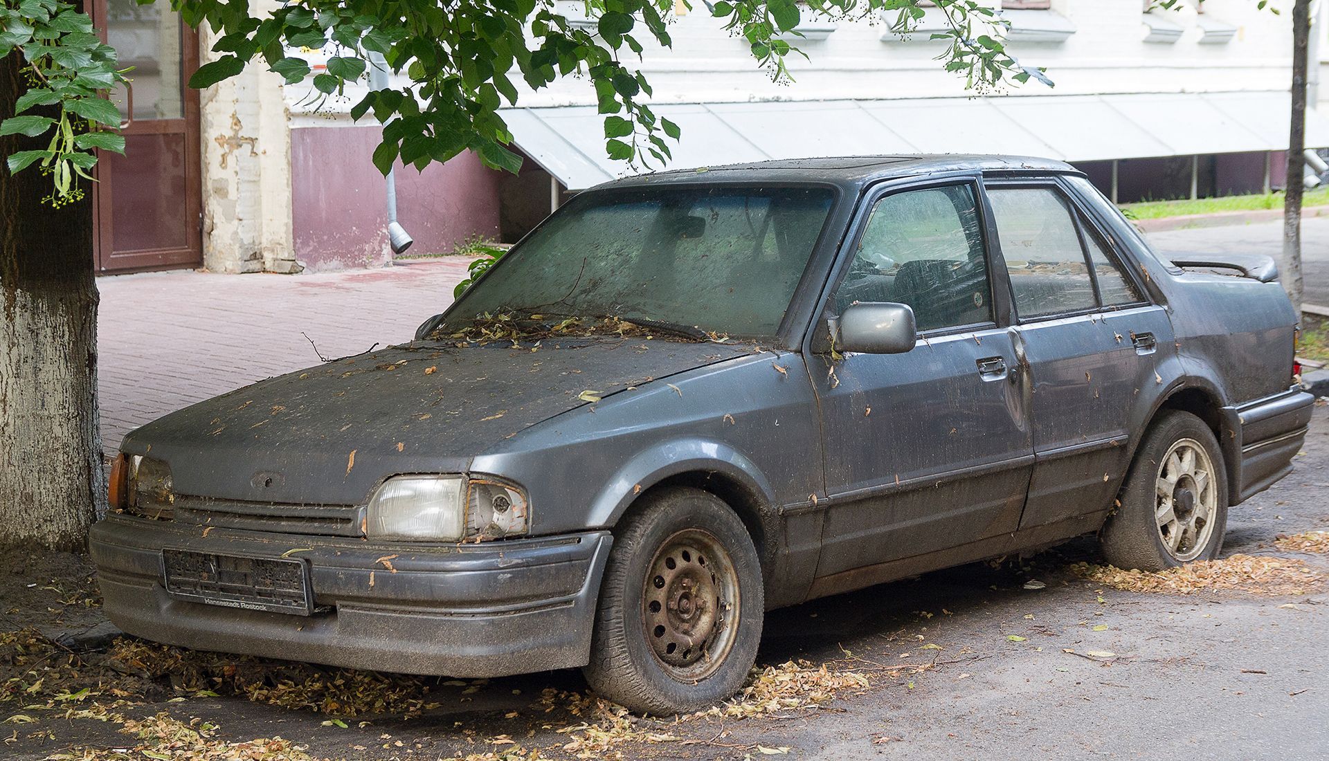Gray, weathered Ford Escort parked on a city street, covered in leaves.