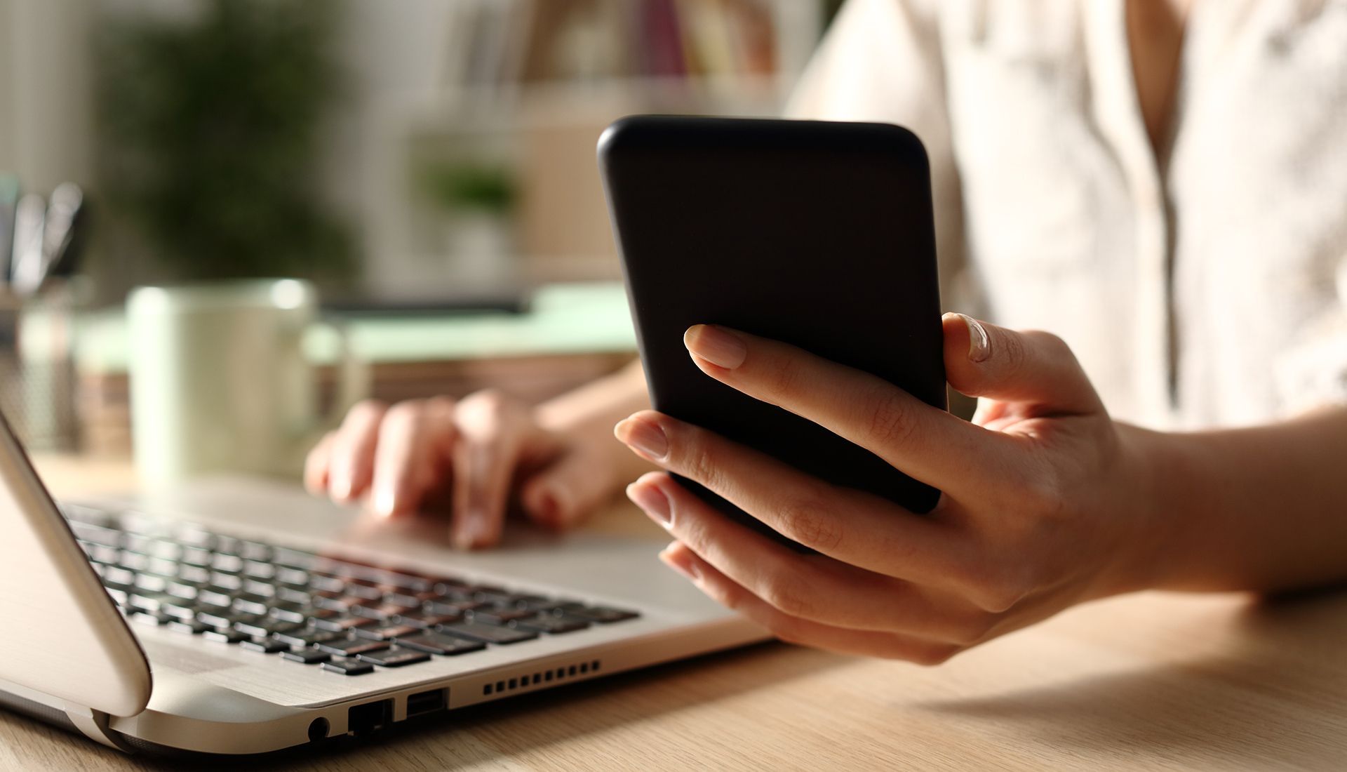Person using a laptop and smartphone at a desk. The focus is on the phone held in the hand.