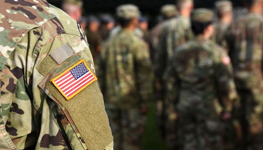 Soldier in camouflage uniform with US flag patch, standing with a group of soldiers outdoors.