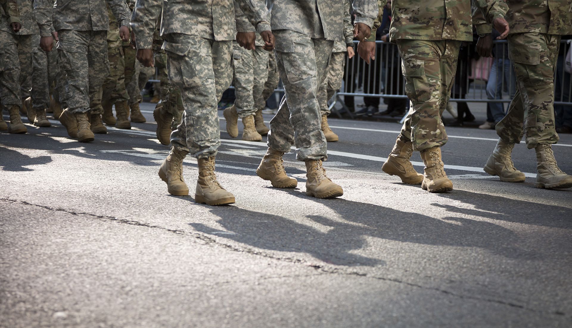 Soldiers in camouflage uniforms marching on a paved street, casting shadows.