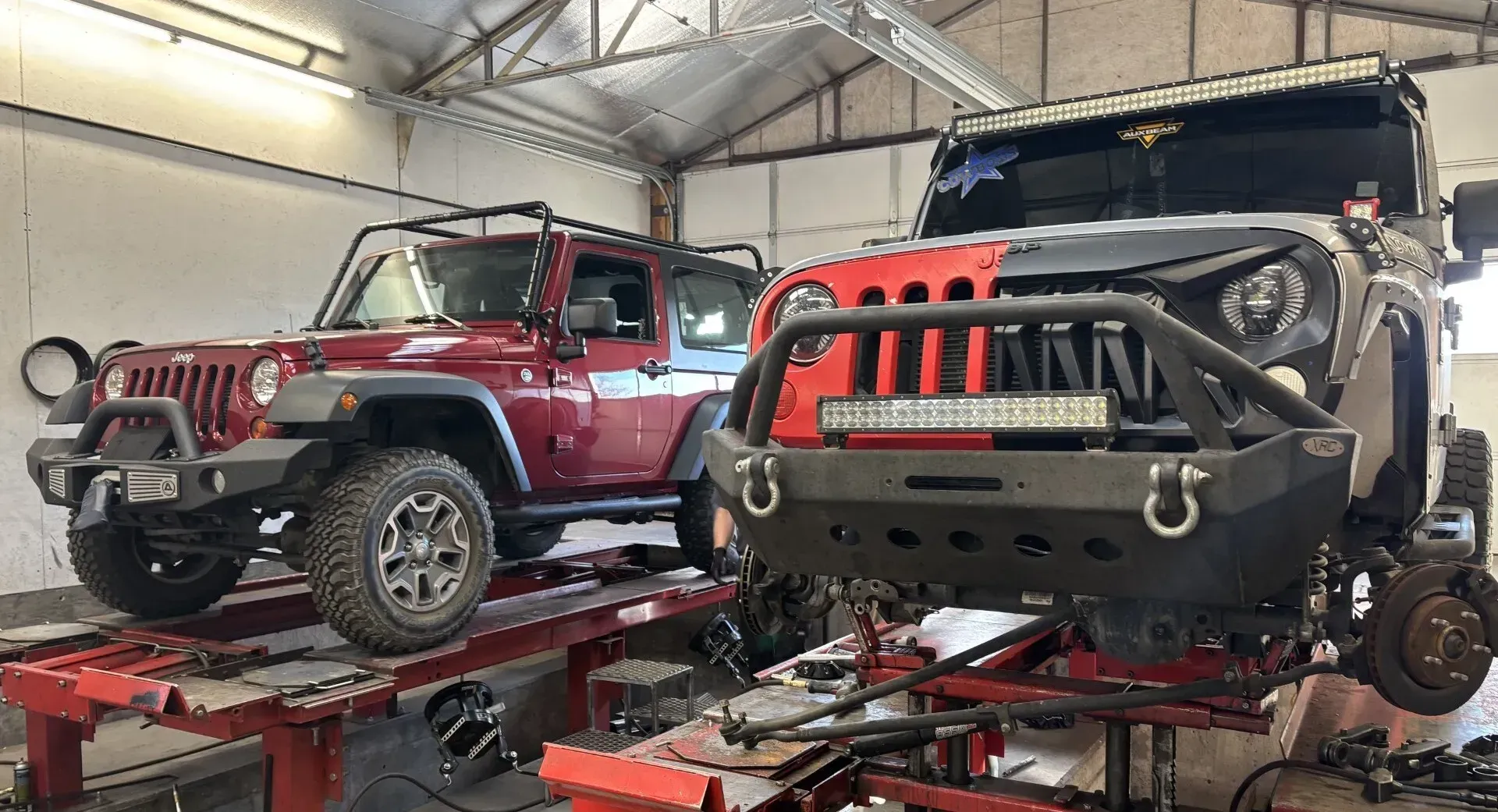 Two modified Jeep Wranglers in a garage, one red and one black, on lifts | Chet's Wheel Alignment