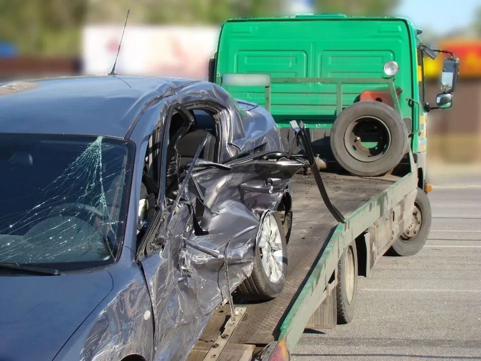 Damaged black car on a tow truck, windshield cracked, driver's side crumpled, tow truck is green.