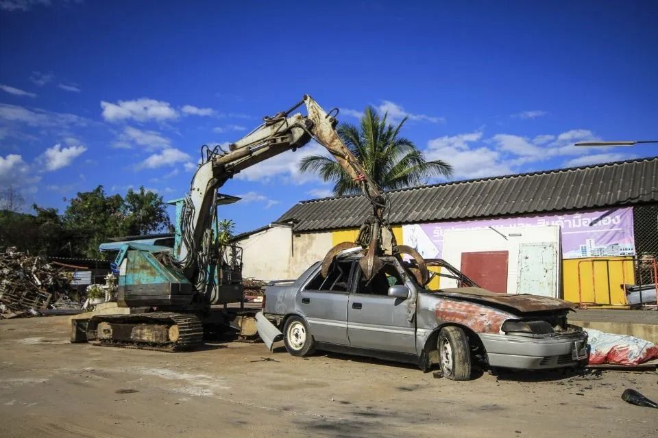 An excavator crushes a damaged gray car in a salvage yard. Blue sky and buildings in the background.