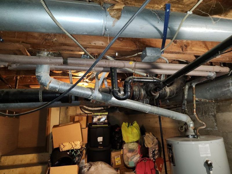 A bunch of pipes are hanging from the ceiling in a basement.
