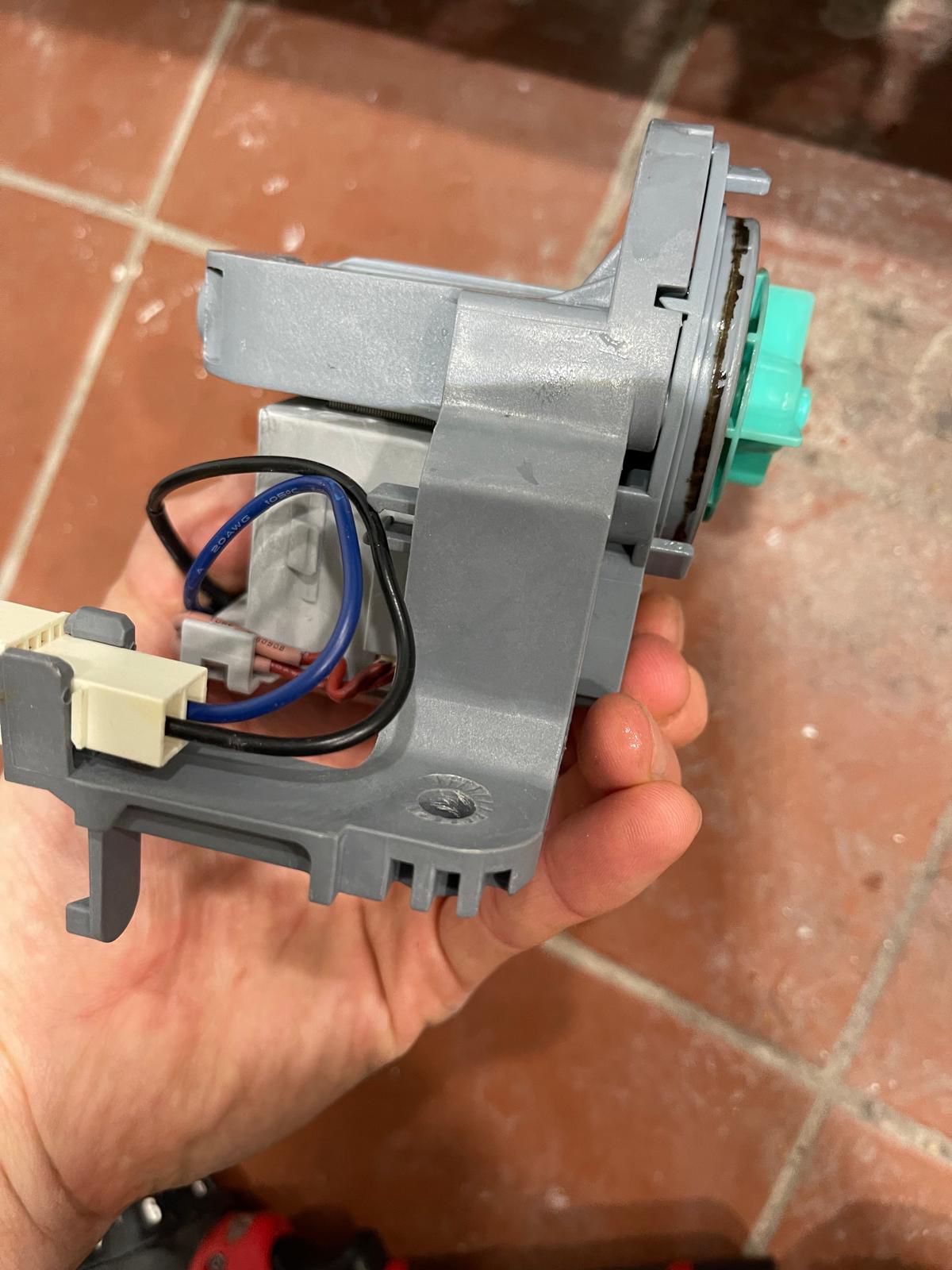 Hand holding a gray and blue dishwasher pump with a green impeller, on a tiled floor.