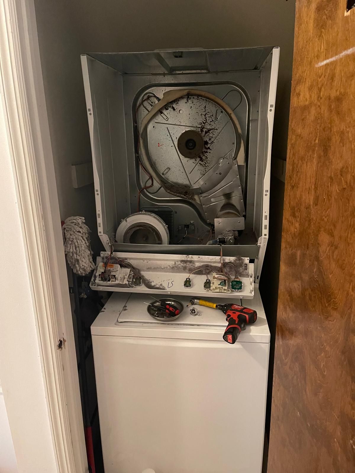 Stacked white washing machine and dryer, dryer's top open, revealing internal components, in a small space.