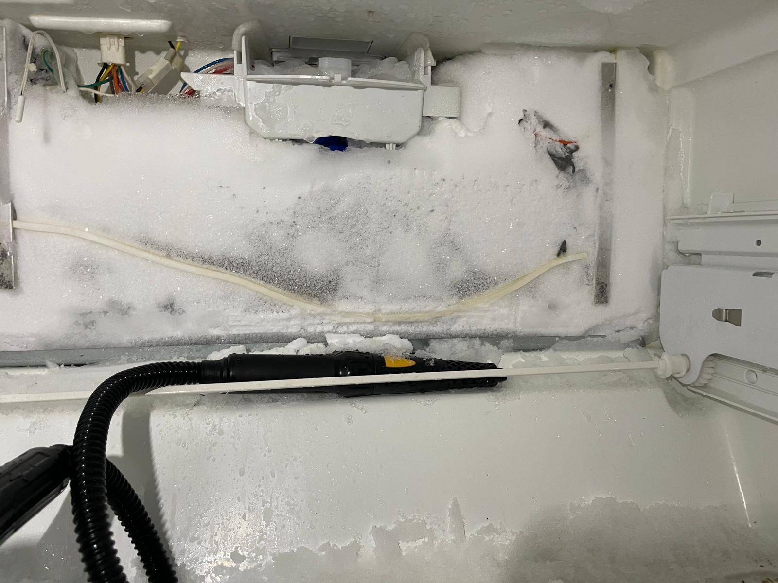 A freezer interior heavily coated in frost. A steam cleaner nozzle is positioned to melt the ice.