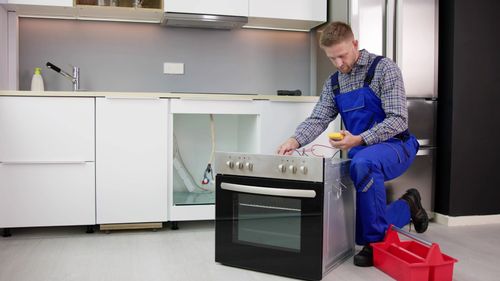A repairman in blue overalls kneels, testing an oven in a white kitchen.