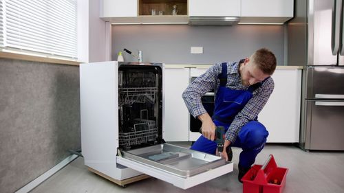 Man in blue overalls repairing a dishwasher in a kitchen.