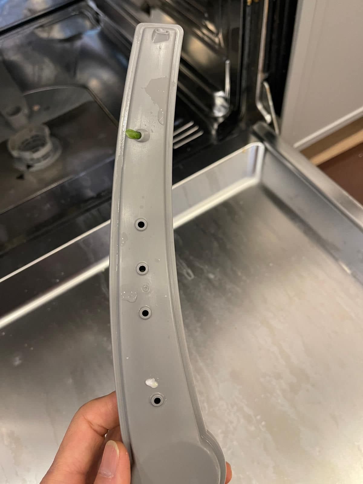 A dishwasher spray arm, gray plastic with several small holes, held in front of an open dishwasher.