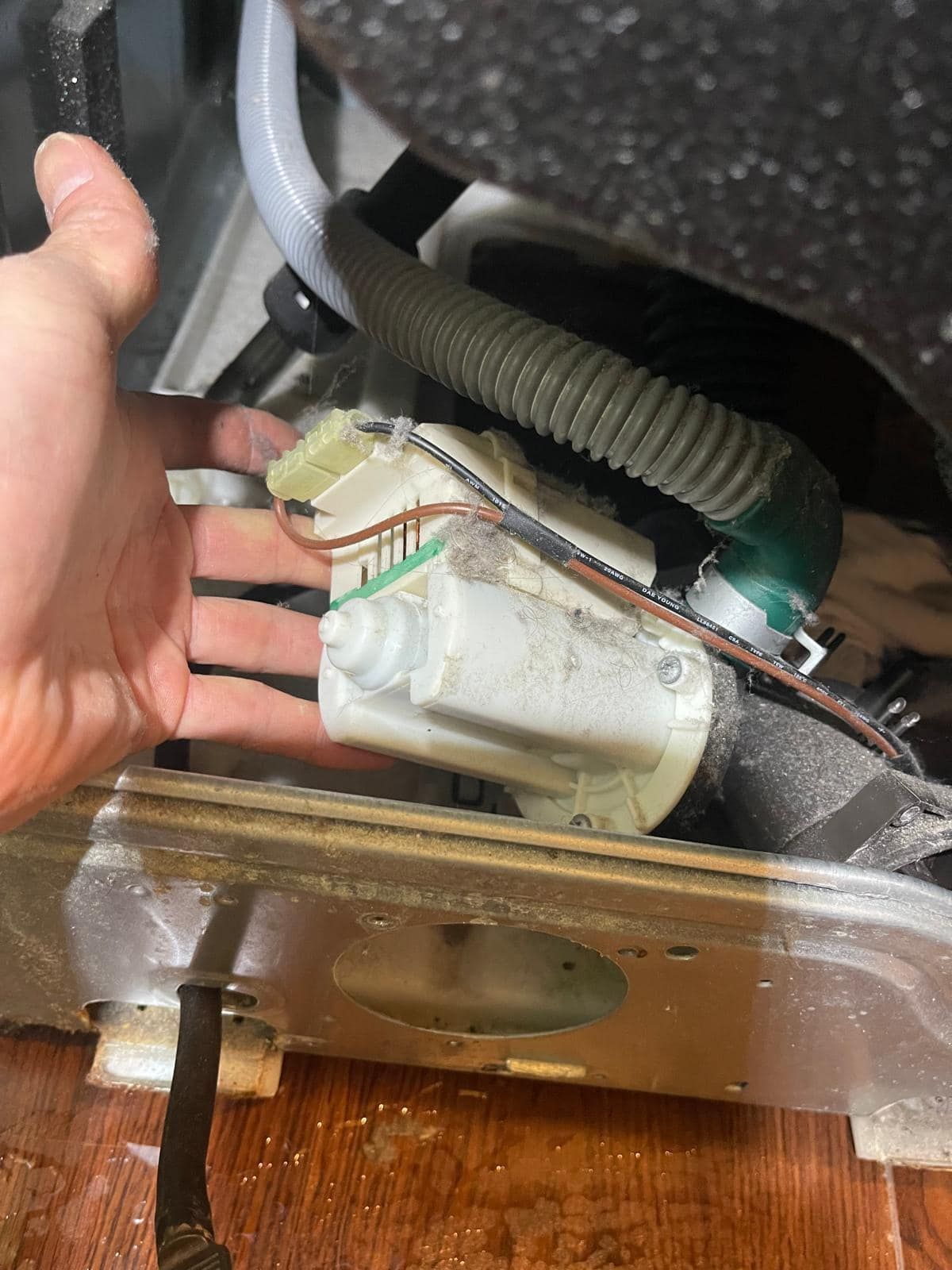 Hand reaching for the drain pump of a washing machine; it is white and dusty, pipes attached.