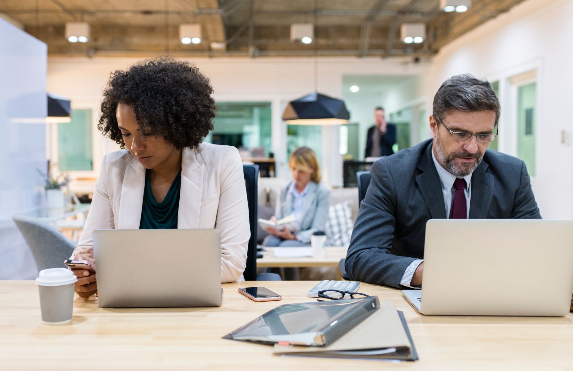 People working on laptops at a shared desk in an office setting.