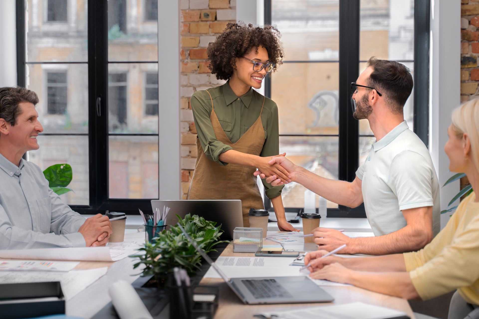 A diverse group in an office, shaking hands, with smiles. Sunlight streams through windows.