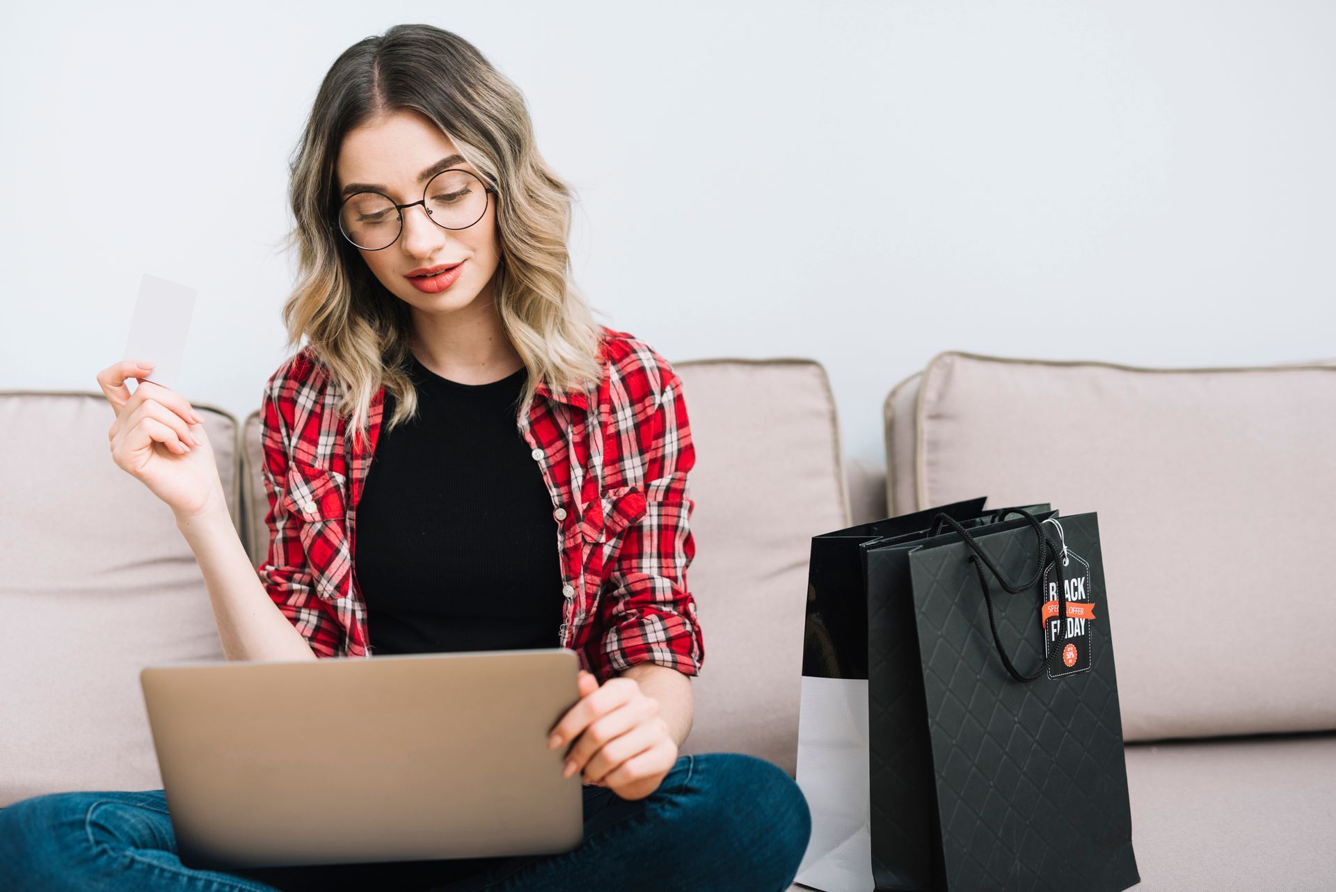 Mulher branca sentada em sofá com notebook no colo e cartão de crédito na mão.