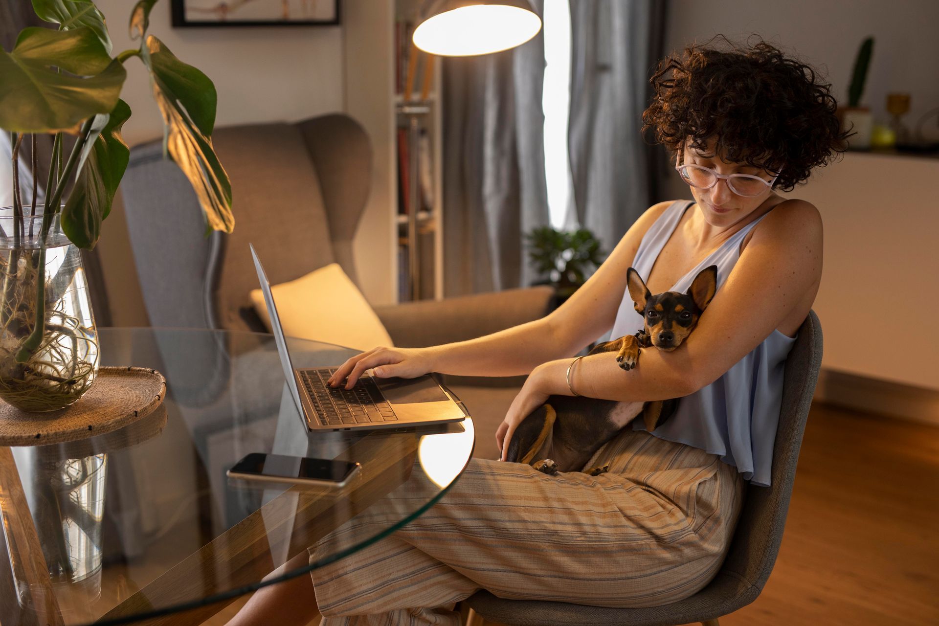 Mulher sentada em frente a uma mesa, com seu notebook apoiado e um cachorro pequeno em seu colo.