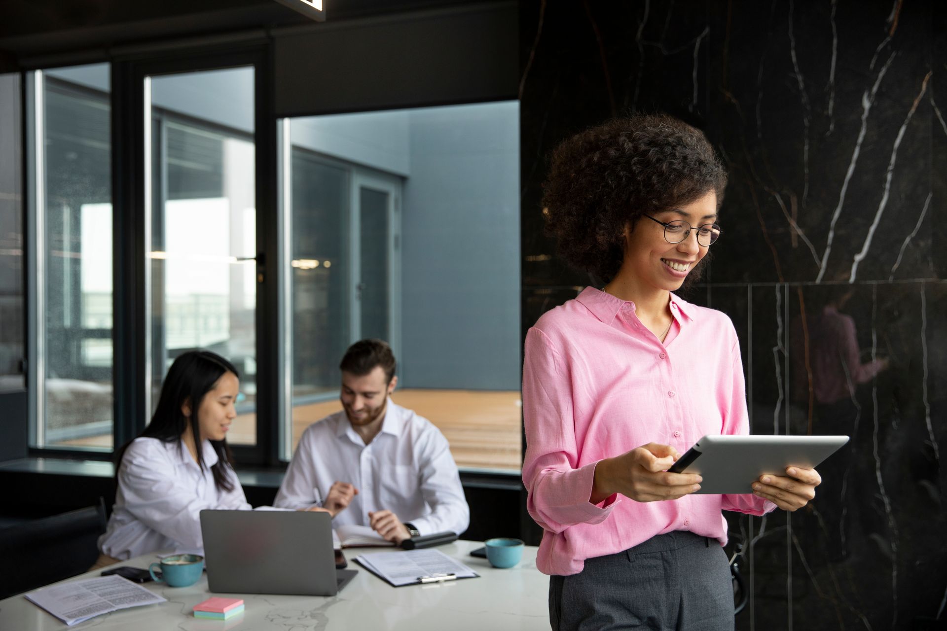 Woman in pink shirt smiles while using tablet, colleagues in background working in office.