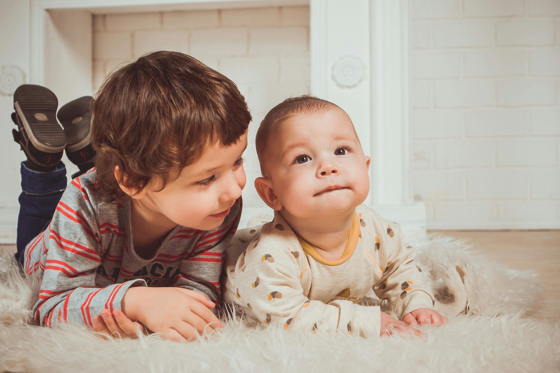 A young boy in striped shirt and baby are laying on the ground  next to each other, on their stomachs. 